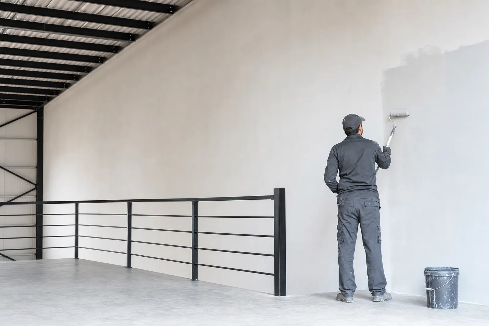 Trabajador pintando un muro interior junto a un barandal de herrería en una nave industrial